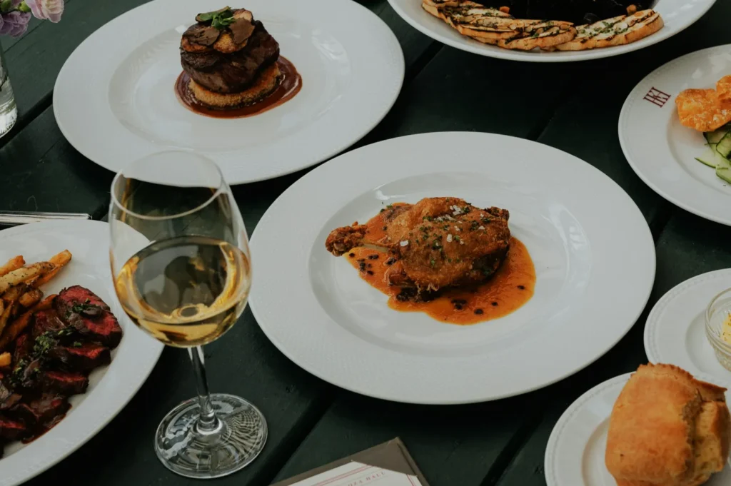 Assorted gourmet dishes on white plates, including steak and fried chicken, surround a glass of white wine on a dark green table, creating an elegant dining scene.