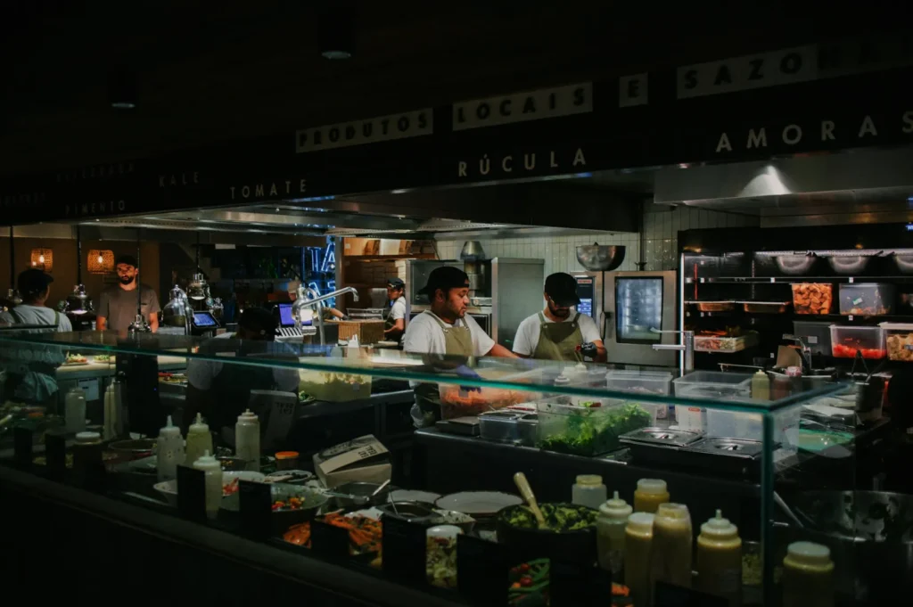 A bustling kitchen with chefs in aprons working behind a glass counter filled with fresh ingredients. The atmosphere is busy and focused.