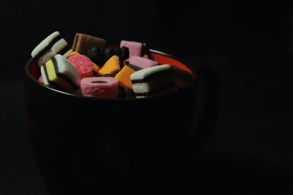 “A dark cup filled with colorful assorted licorice candy, including pink, orange, and striped pieces, against a black background. The mood is playful.”