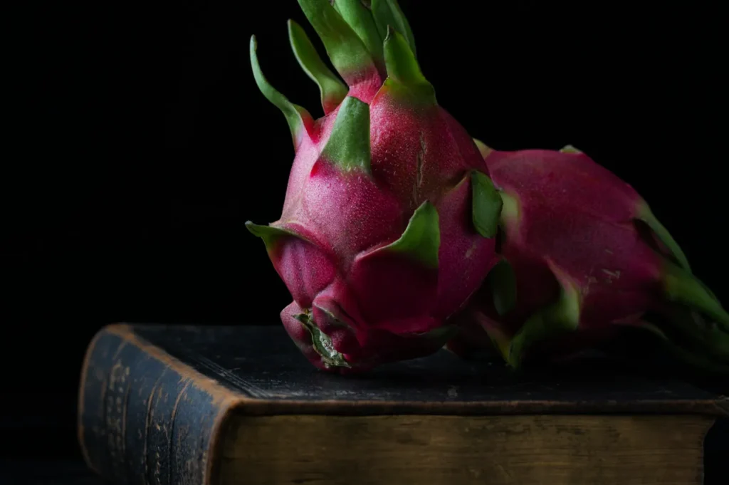 “Two vibrant pink dragon fruits with green-tipped scales rest on an antique book against a dark background, creating a serene, artistic still life.”