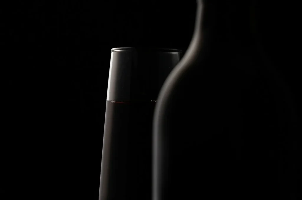 A glass of red wine and an elegant bottle cast in shadow against a black background, creating a mysterious and sophisticated ambiance.