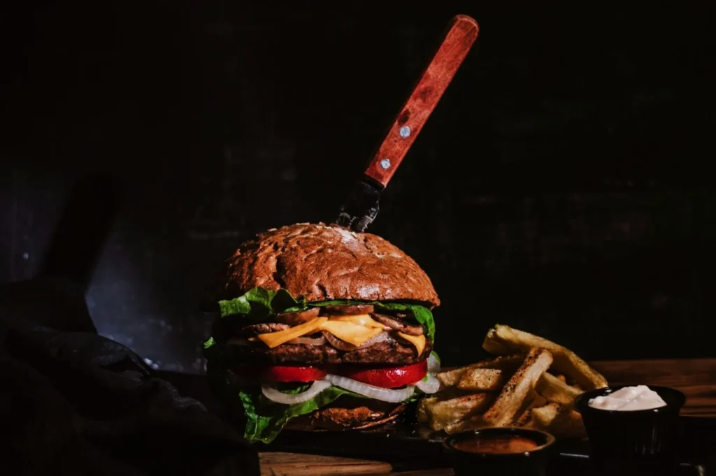 “A large gourmet burger with lettuce, cheese, tomatoes, and onions has a knife through the bun. It is paired with seasoned fries and two dips on a dark background.”