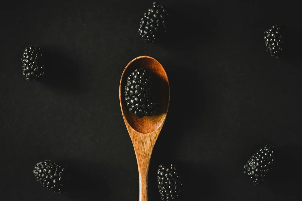 “A wooden spoon holds a single blackberry, surrounded by several more on a dark background. The contrast highlights the rich, textured surface of the berries.”