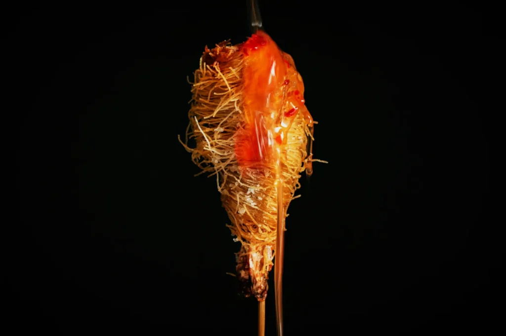 “A close-up of a deep-fried shrimp wrapped in crispy noodles, coated with a glossy, reddish-orange sauce, set against a dark background. The texture is intricate and appetizing.”