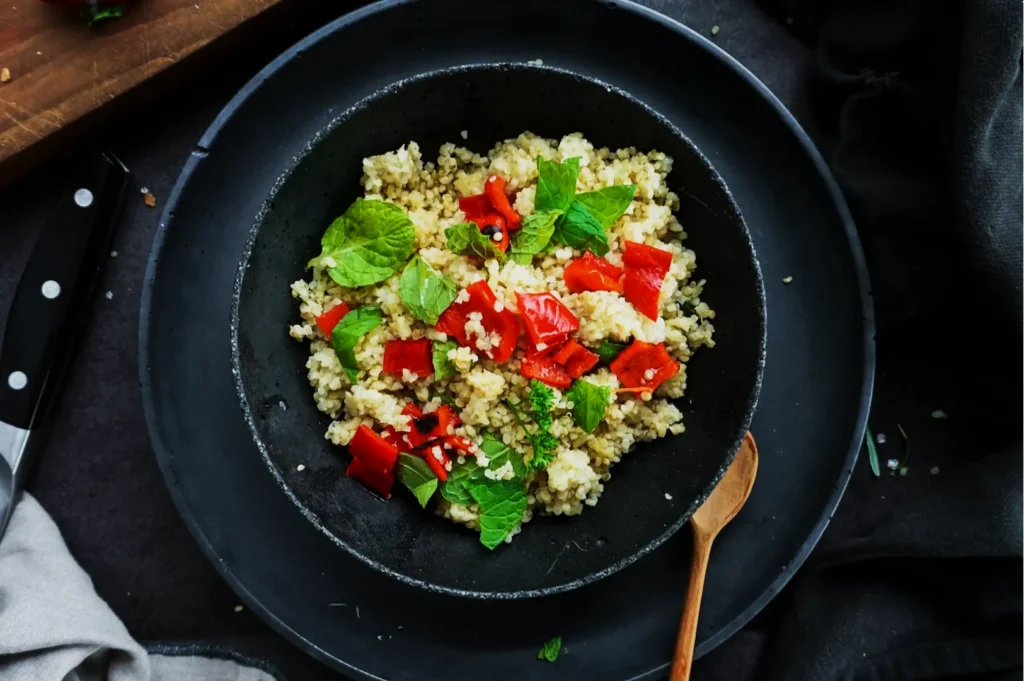 “A black bowl on a dark surface filled with quinoa, topped with fresh mint leaves and chopped red bell peppers, next to a wooden spoon.”