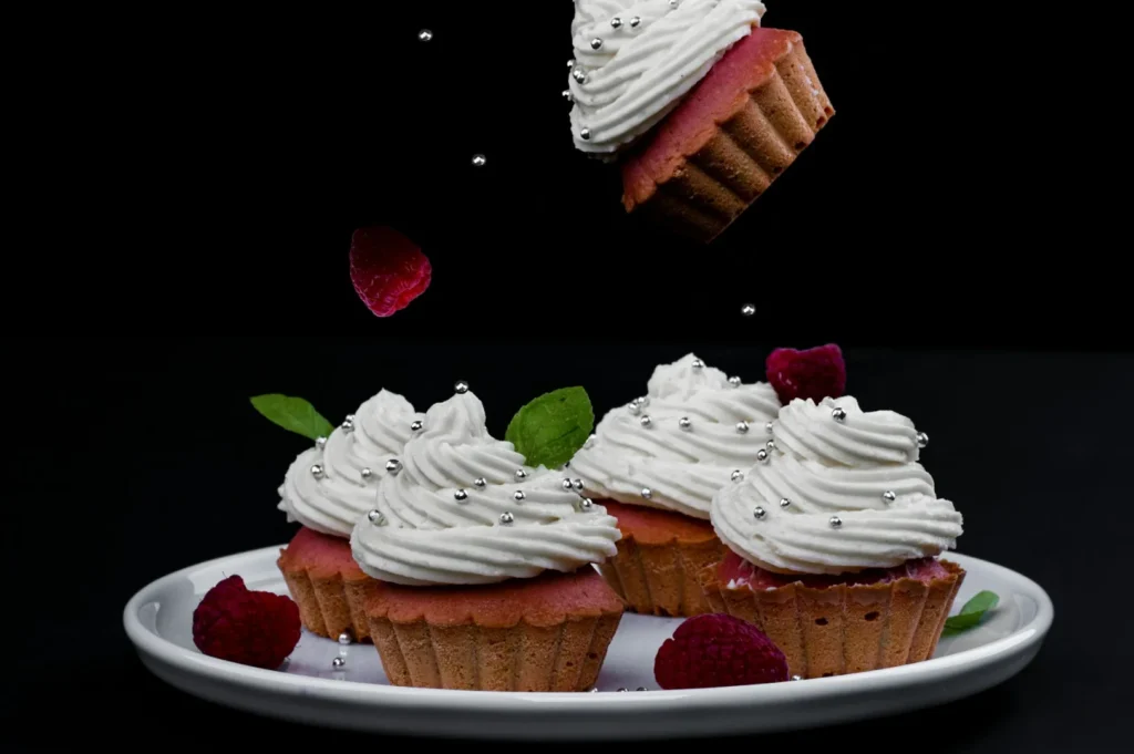 “Plate of cupcakes with white frosting, silver dragees, raspberries, and mint leaves. One cupcake drops from above, against a black background.”