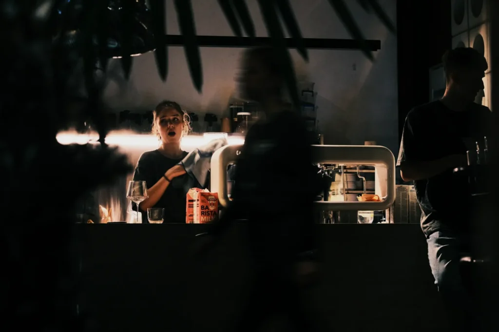 Barista in a dimly lit café cleans glassware behind the counter. Two blurred figures pass by, creating a bustling yet intimate ambiance.