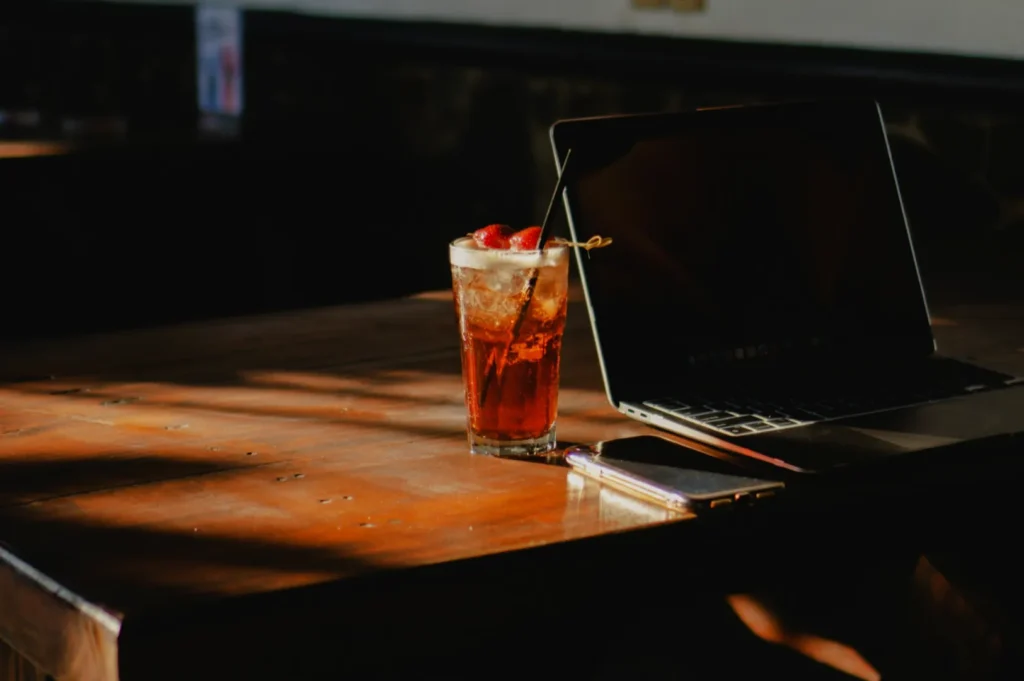 A dimly lit wooden table with a laptop and smartphone. A glass of iced tea with a straw and garnished with raspberries is bathed in soft sunlight, creating a relaxed atmosphere.
