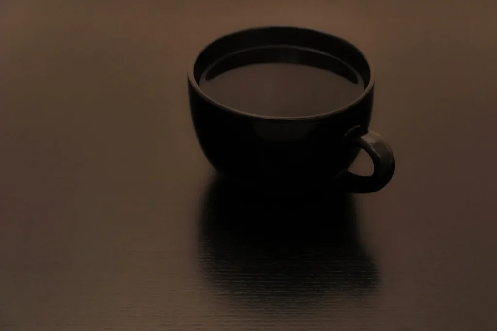 “A black coffee cup filled with dark liquid sits on a smooth, dark wooden table. The minimalist setting conveys a sense of calm and simplicity.”