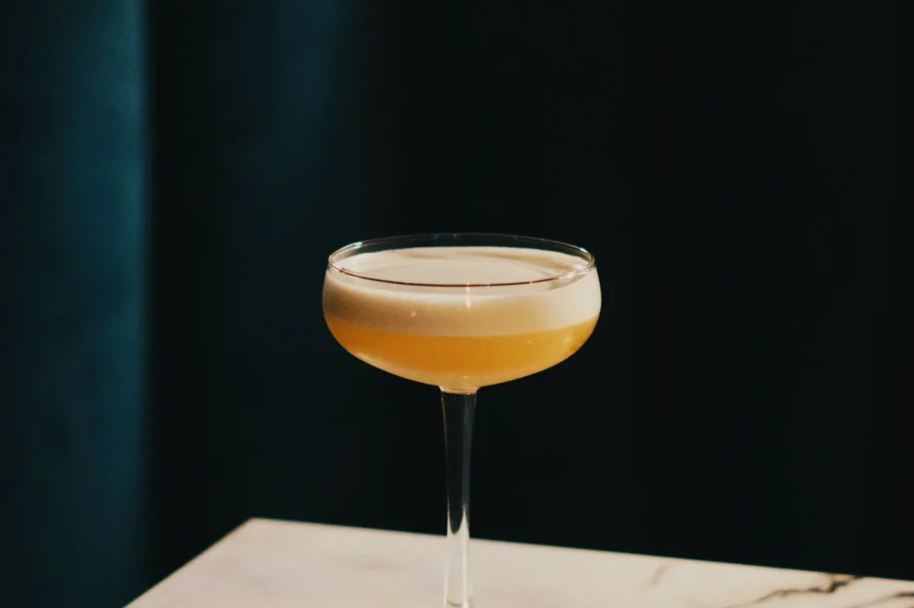 A frothy cocktail in a coupe glass sits on a marble surface. The drink is golden, with a foamy top, set against a dark, moody background.