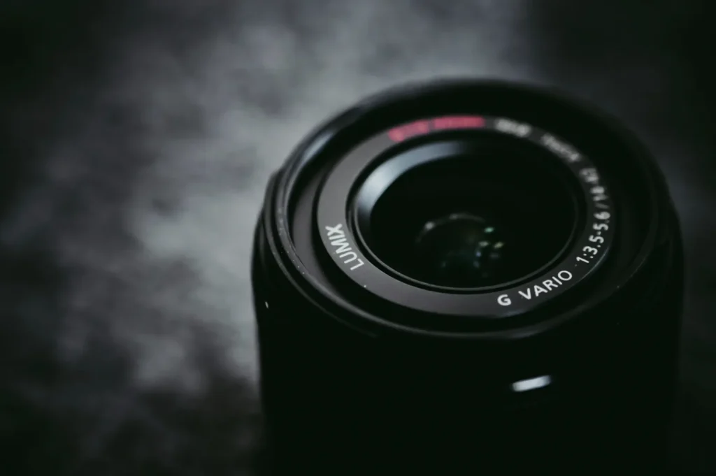 “Close-up of a dark camera lens with "Lumix" and "G Vario" inscriptions. The background is softly blurred, creating a dramatic and focused effect.”