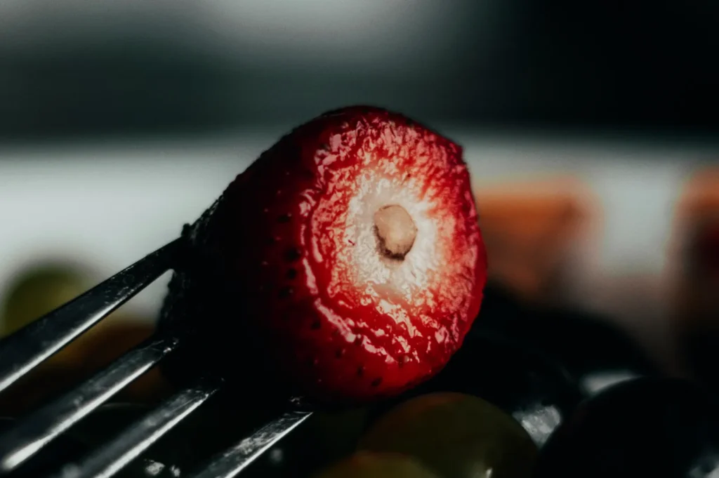 “Close-up of a strawberry half skewered on a fork, with a shiny, red surface. Blurry dark and light fruits are visible in the background, adding contrast.”