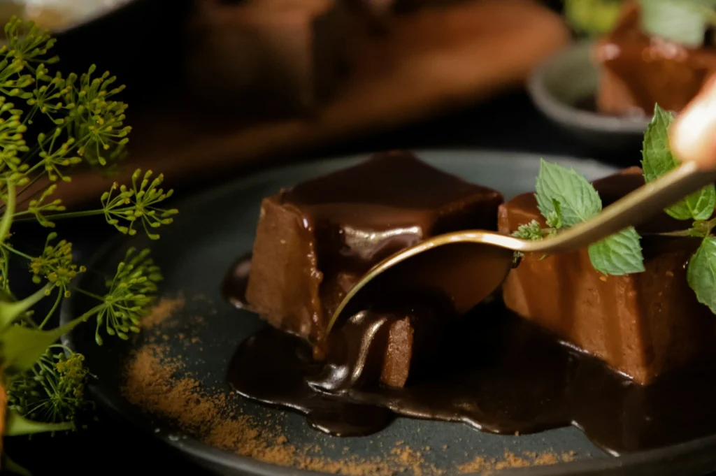 “Close-up of rich chocolate cake squares on a gray plate, covered in glossy chocolate sauce. A spoon cuts in, with green garnishes beside.”