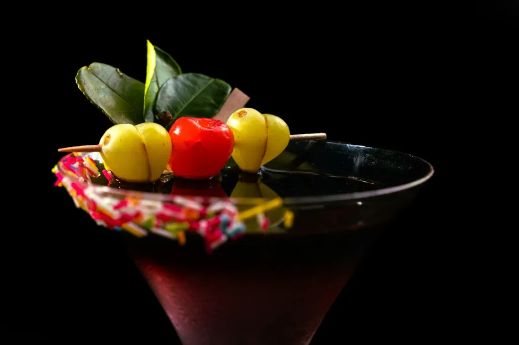 A vibrant cocktail in a glass, garnished with a skewer of cherries and green leaves. The rim is decorated with colorful sprinkles, set against a black background.