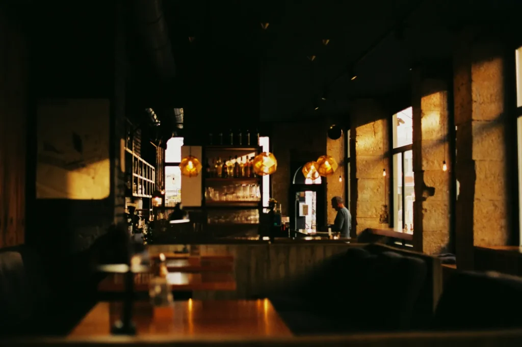 Cozy cafe interior with dim lighting and golden pendant lamps. Wooden tables and textured walls create a warm, inviting ambiance. A person stands at the bar.