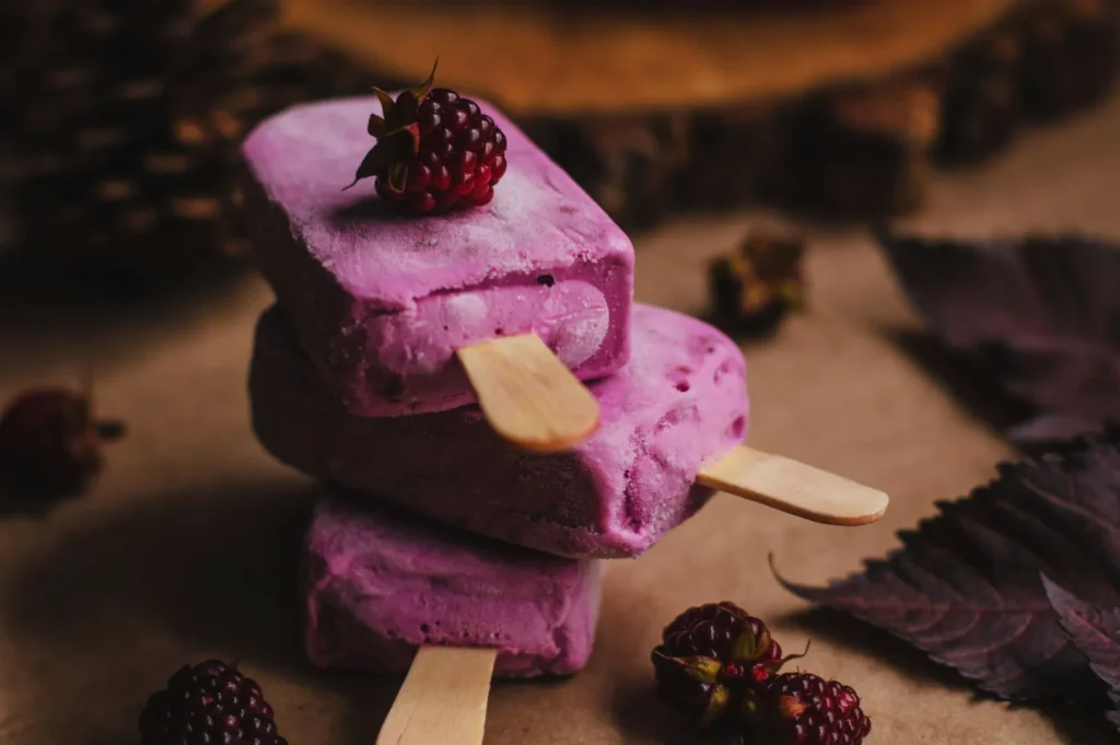 Three stacked pink berry popsicles with wooden sticks, topped with fresh blackberries. Rustic background with leaves and warm lighting.