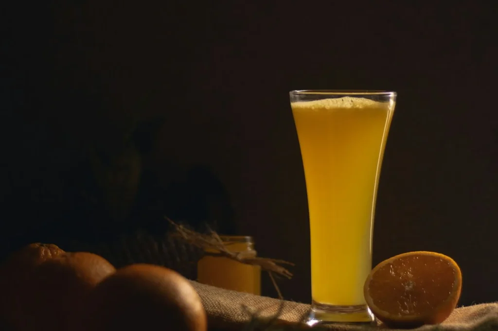 “A tall glass of orange juice is on a table with dim lighting. Whole and half-cut oranges sit beside it, creating a warm, inviting atmosphere.”