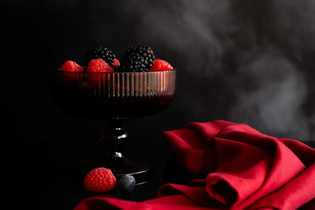 Dark glass bowl with blackberries and raspberries on a black surface, beside a vibrant red cloth. The mood is elegant and dramatic.