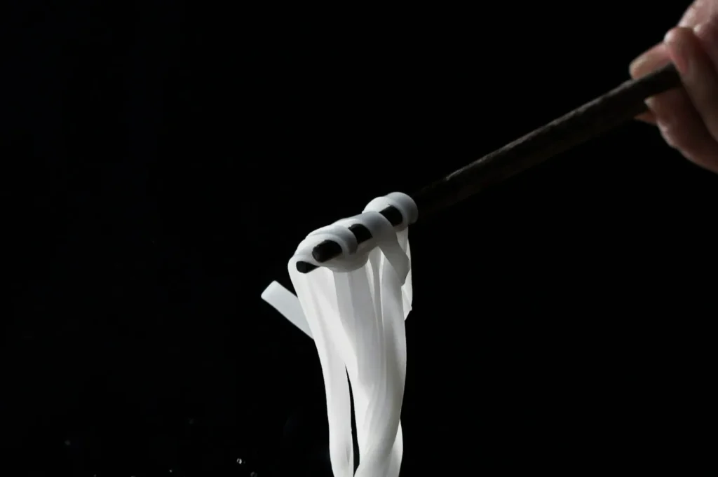 “Close-up of chopsticks holding thin, white noodles against a black background. The image conveys a minimalist and elegant culinary scene.”
