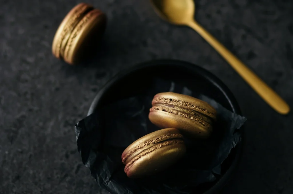 “Golden macarons in a black bowl with crumpled paper, placed on a dark surface. A gold spoon lies nearby, creating an elegant, luxurious feel.”