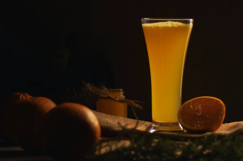 “A tall glass of frothy orange juice sits on a table with whole and halved oranges nearby, warmly lit against a dark background, conveying a fresh, inviting feel.”
