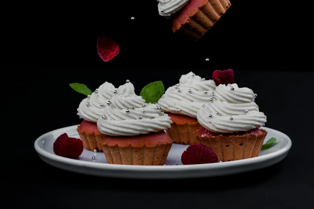 “Four cupcakes with white frosting and silver sprinkles on a white plate. Surrounded by raspberries and mint leaves, one cupcake is falling. Black background.”