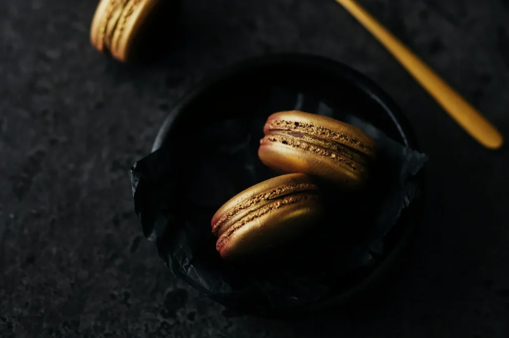 “Gold macarons on dark tissue paper in a black bowl, with a matte gray surface and a gold spoon nearby, creating a luxurious and elegant feel.”