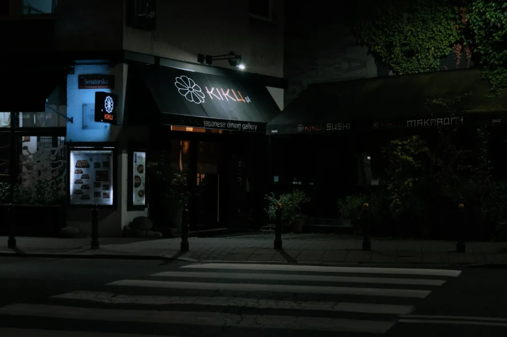 Dimly lit Japanese restaurant exterior at night, with a black awning and glowing "KIKU" sign. Quiet, serene street with a crosswalk.