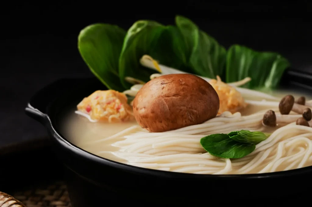 A black bowl of creamy noodle soup with a mushroom, leafy greens, and small meatballs. Fresh and comforting with earthy tones.