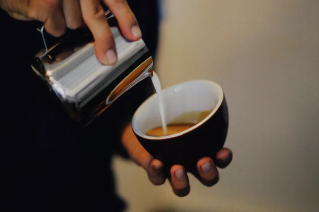 “A person is pouring steamed milk from a metal pitcher into a cup of coffee, creating latte art. The background is blurred, emphasizing the creamy texture.”
