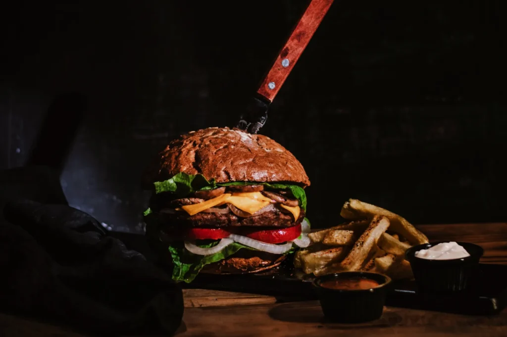 A rustic burger with cheese, lettuce, and tomato is skewered by a large knife. It sits beside crispy fries with dipping sauces in dim lighting.