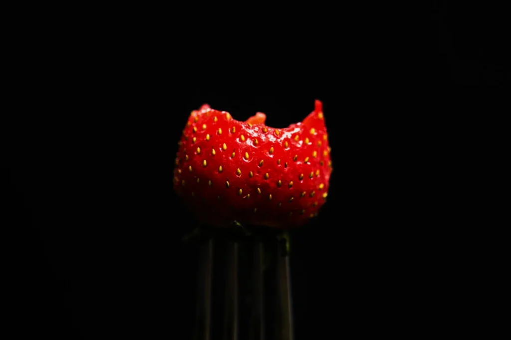 “A vibrant red strawberry with a bite taken out is perched on a fork against a dark background, highlighting its freshness and texture.”