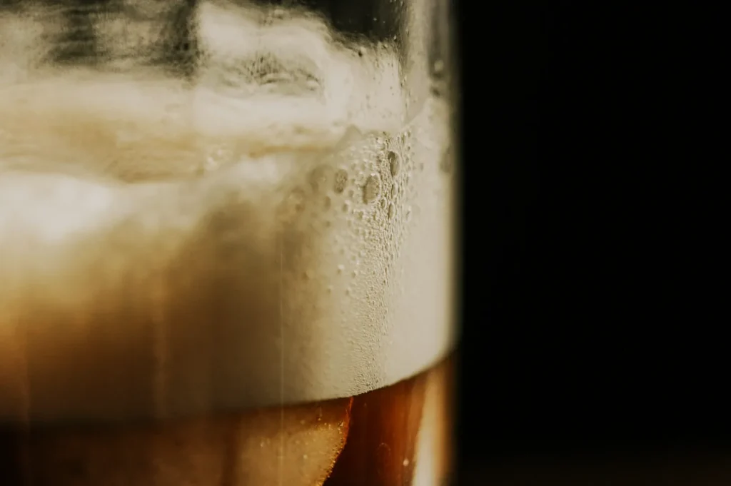 “Close-up of a frothy beer in a glass with a dark background. The foam is prominent, with bubbles visible, creating a refreshing, inviting tone.”