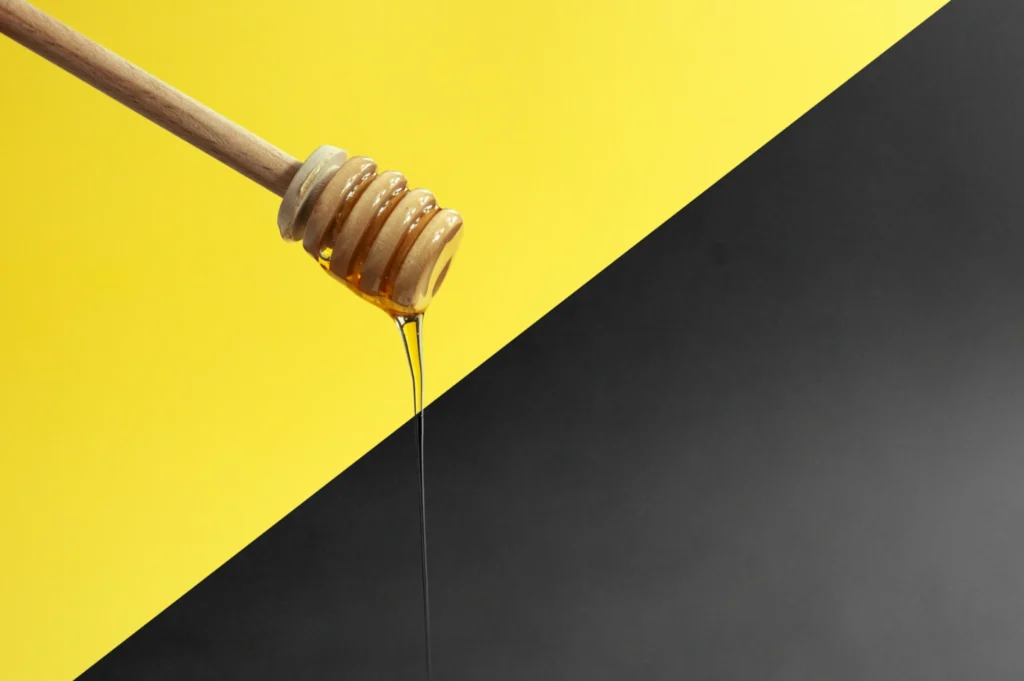 “A honey dipper drips honey diagonally against a background split into bright yellow and dark gray halves, creating a striking contrast.”