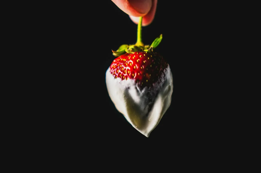 “A hand holds a ripe strawberry dipped in smooth white chocolate against a dark background. The contrast highlights the strawberry’s vibrant red color.”