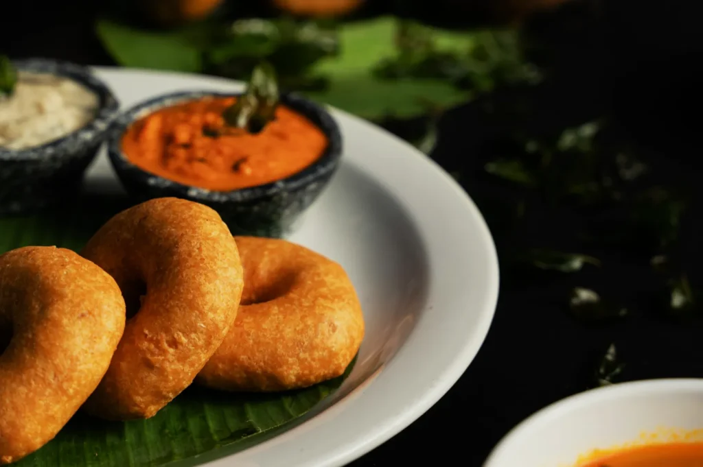 Three crispy Indian vadas on a white plate lined with a banana leaf, served with orange and white chutneys, creating a delicious and appetizing scene.