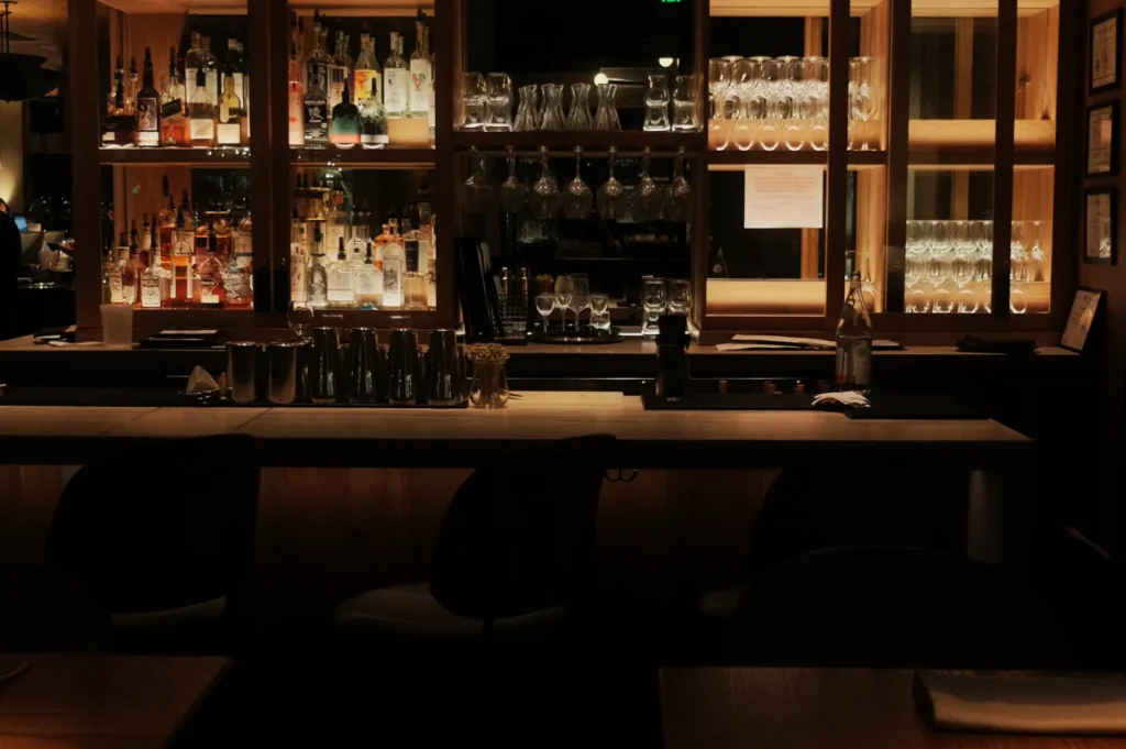 Cozy bar interior with a softly lit wooden bar counter, stools, an array of liquor bottles, and hanging glasses, creating a warm, inviting ambiance.