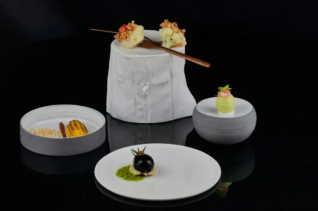 “A sophisticated culinary display featuring artfully arranged small dishes on a dark reflective surface. Includes a white stand with skewers and elegantly plated bites.”