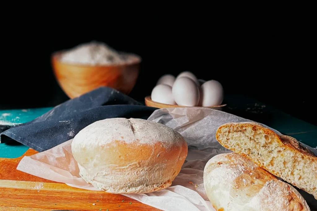 “A close-up of freshly baked, flour-dusted bread on parchment paper, with eggs and a bowl of flour in the background. The scene conveys a rustic, homemade feel.”