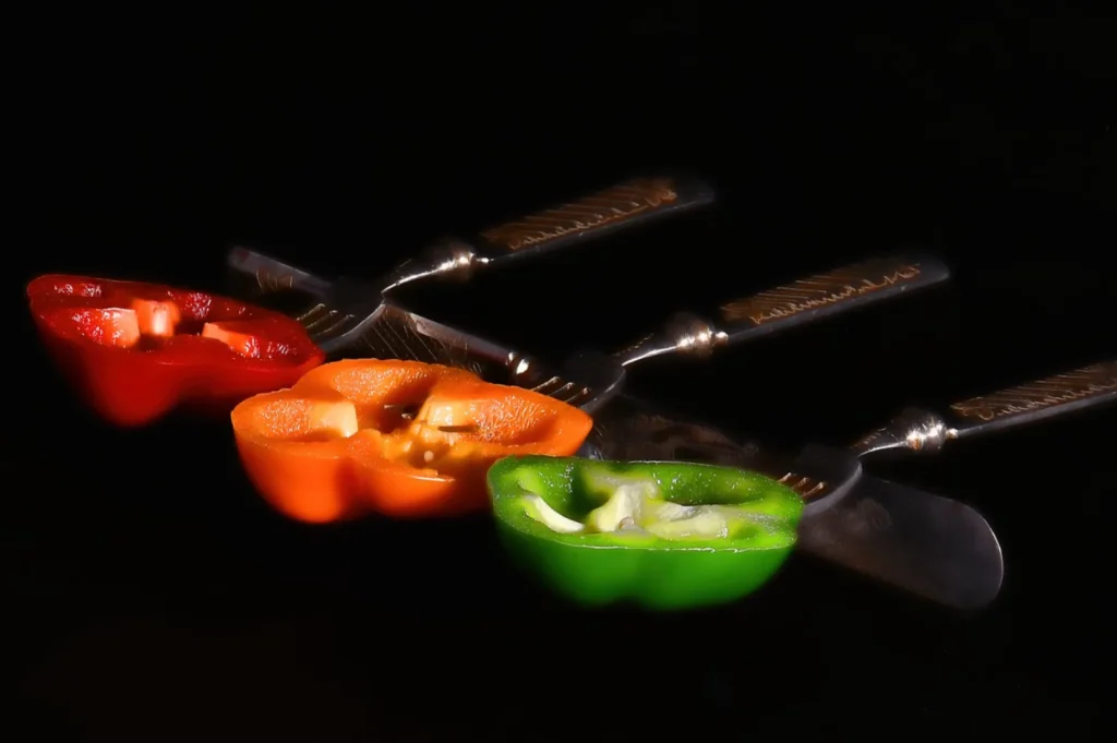 Three colorful bell pepper halves—red, orange, and green—are pierced by parallel silver forks against a dark background, creating a vibrant and playful composition.