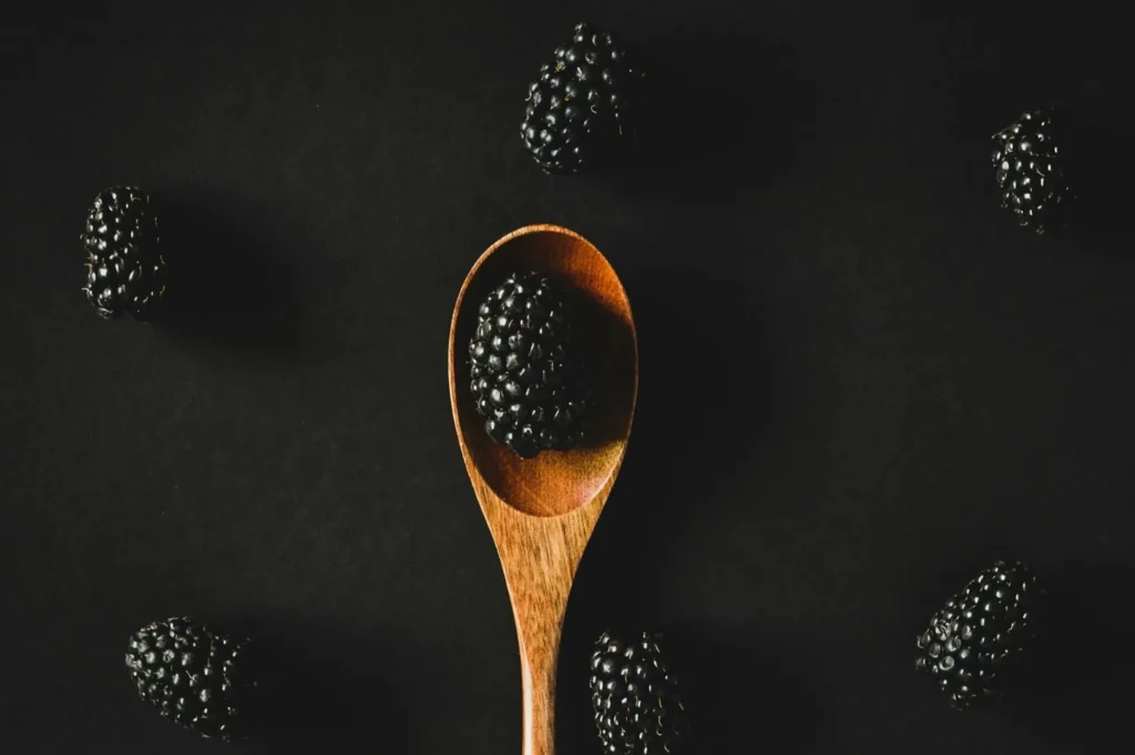 “A wooden spoon holds a blackberry against a dark background, surrounded by scattered blackberries, creating a minimalist and elegant composition.”