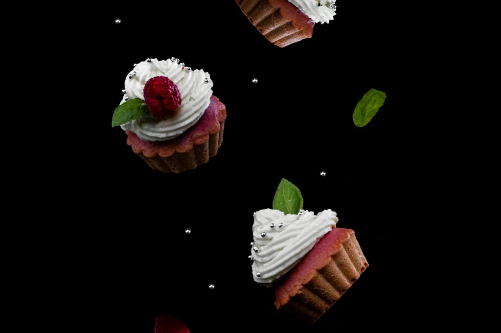 “Three cupcakes with white frosting, raspberries, and mint leaves float against a black background, surrounded by tiny silver balls, creating a whimsical and elegant scene.”