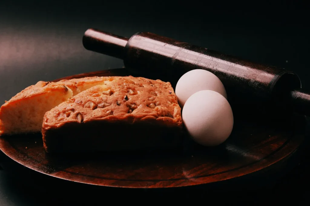 “A wooden board holds a slice of nutty bread, two white eggs, and a dark rolling pin, set against a moody dark background.”