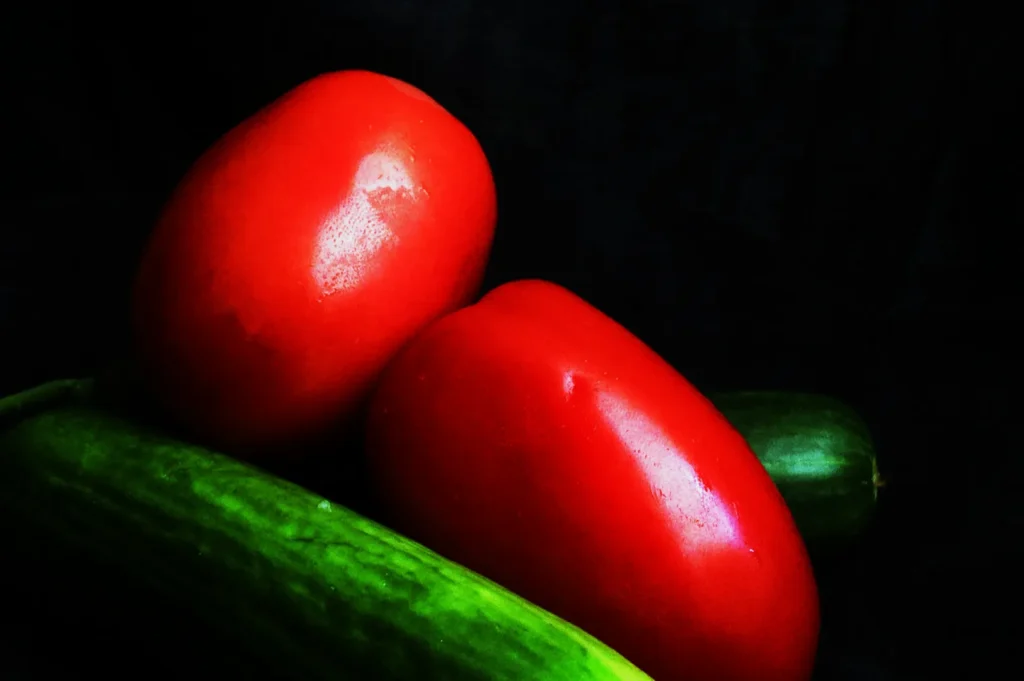 “Two vibrant red tomatoes rest atop a shiny green cucumber against a dark background, creating a contrast that highlights their freshness and color.”