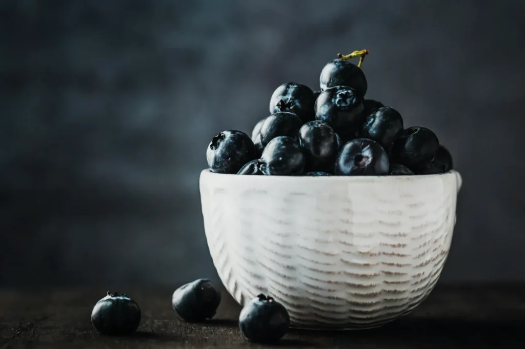 “A white ceramic bowl filled with fresh blueberries sits on a dark wooden surface, set against a blurred gray background, conveying a simple and rustic vibe.”