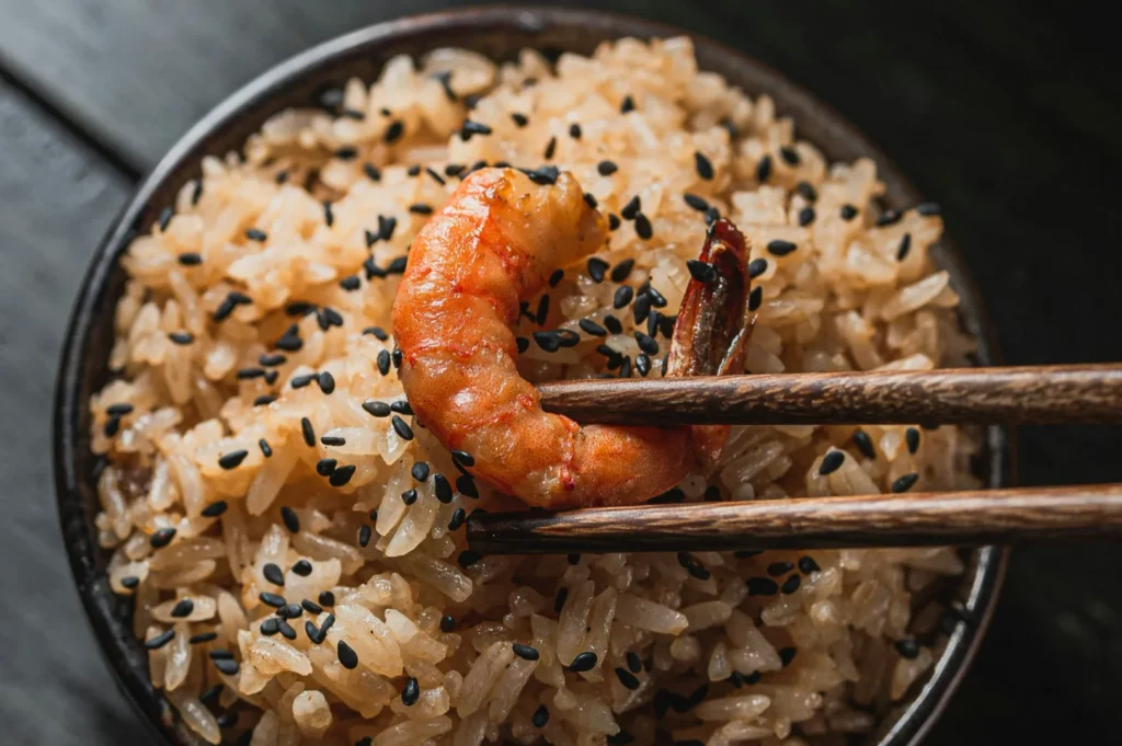 “A bowl of rice garnished with black sesame seeds is topped with a shrimp held by wooden chopsticks. The dish has a savory, appetizing appearance.”