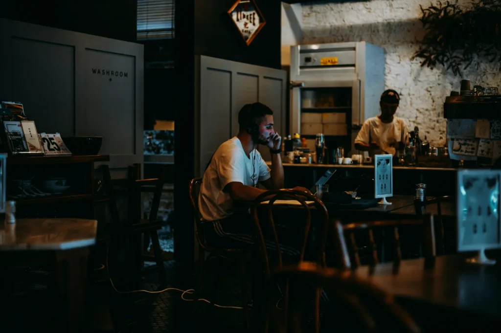 A man with headphones sits at a dimly lit cafe table, focused on a laptop. Another person works behind the counter. Cozy, productive atmosphere.