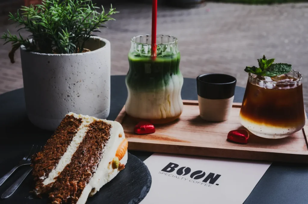 Carrot cake slice and layered green tea drink on a table with a wooden tray, featuring an iced coffee and small plant, creating a cozy café vibe.