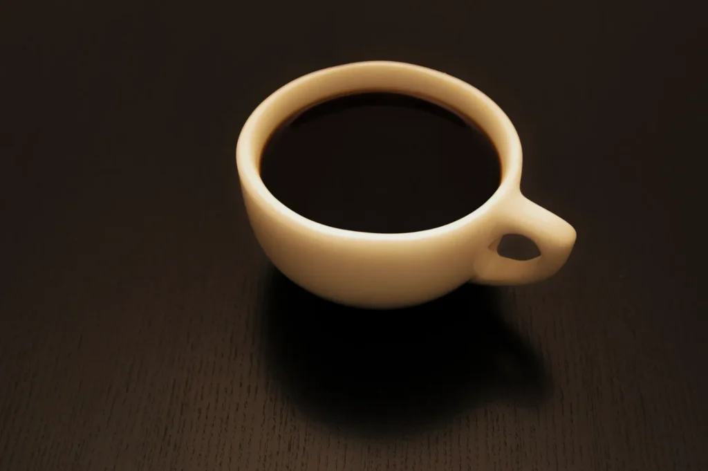 “A small white cup filled with dark black coffee sits on a smooth, dark wood table. The soft lighting creates a warm and inviting atmosphere.”