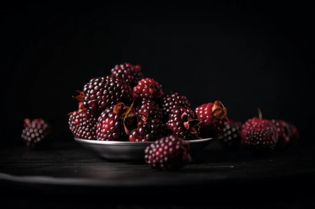 A small plate filled with fresh, dark blackberries sits on a dark surface against a black background, creating a rich, moody ambiance.